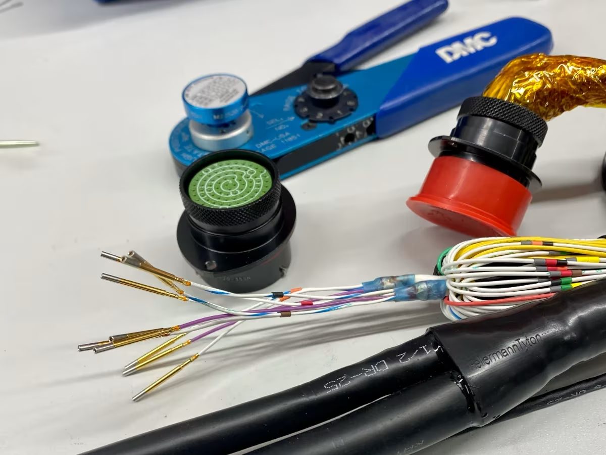Faisceau motorsport en cours de réalisation chez O-ONE : connecteur circulaire à insert vert, contacts dorés sertis, gaine DR-25 et pince à sertir DMC