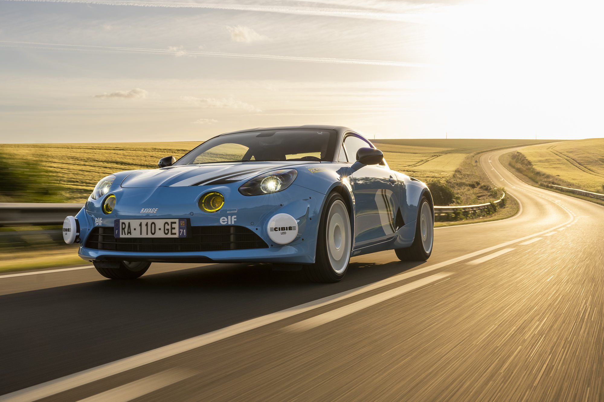 Alpine A110 Ravage Groupe 4 bleue en décoration San Remo, phares longue portée Cibié, sur une route de campagne au soleil couchant